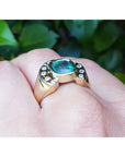 Gold ring with a natural emerald on a hand against a blurred green leafy background.