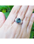 Hand wearing a white gold ring with a green bluish natural sapphire against a green leafy background