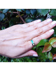 A hand wearing a men's yellow gold emerald ring against a green leafy background.