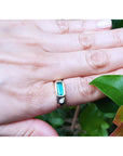 A person's hand wearing a mens emerald ring in yellow gold on a leafy background.