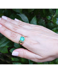 Hand wearing a gold ring for men with a natural emerald against a green leafy background.