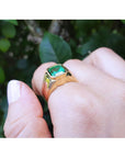 Gold men's ring with a natural emerald on a hand against a blurred green leafy background.