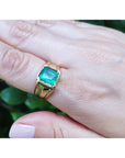Gold ring for man with a square natural emerald on a hand against a blurred green background.