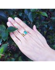Hand wearing a gold men's emerald ring with accent diamonds against a leafy background.