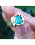 Gold ring with a natural emerald held between fingers against a blurred natural background.
