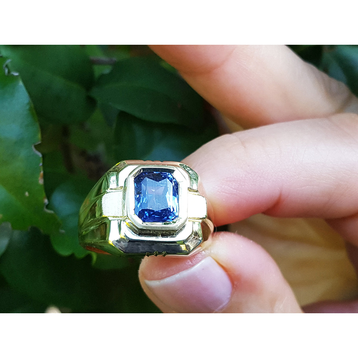 Gold ring with a blue natural sapphire gemstone held between fingers against a green leafy background