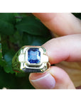 Gold ring with a blue natural sapphire gemstone held between fingers against a green leafy background