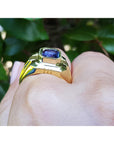 Gold ring with a blue natural sapphire sagemstone on a finger against a blurred green leafy background