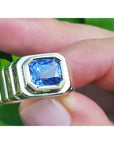 Yellow gold ring with a blue bezel set sapphire gemstone held between fingers against a green leafy background