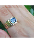 Gold ring with a blue sapphire gemstone on a hand against a blurred green background