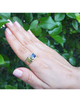Hand wearing a gold ring with a blue natural sapphire gemstone against a green leafy background