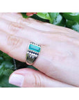 A yellow gold ring with an emerald on a hand against a green leafy background.