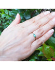 Hand wearing a yellow gold minimalist emerald ring against a green leafy background.