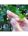 Hand holding a natural minimalist solitaire emerald ring against a blurred green leafy background