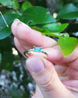 Hand holding a natural minimalist solitaire emerald ring against a blurred green leafy background
