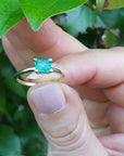 A minimalist emerald solitaire ring held between fingers against a green leafy background.