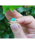Minimalist 14K yellow gold ring with a square-cut natural Colombian emerald, held by fingers near green leaves.