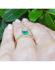 A solitaire emerald gold ring on a hand against a leafy background.