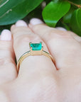 A solitaire emerald gold ring on a hand against a leafy background.