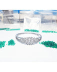 White gold bangle bracelet with diamonds, surrounded by loose green Colombian emeralds and small bags.