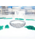 White gold bangle bracelet with diamonds, surrounded by loose green Colombian emeralds and small bags.