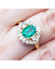 A cushion cut Muzo colombian emerald surrounded by a halo diamond 14k gold ring on a person's finger.