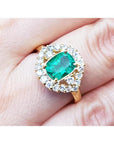 A cushion cut Muzo colombian emerald surrounded by a halo diamond 14k gold ring on a person's finger.