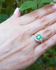 Hand wearing an emerald ring with a green leafy background.