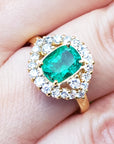 A cushion cut Muzo colombian emerald surrounded by a halo diamonds ring on a person's finger.