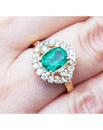 A cushion cut Muzo colombian emerald surrounded by a halo diamonds ring on a person's finger.