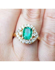 A cushion cut natural Muzo emerald ring with a halo diamond on a finger.