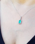An emerald-cut emerald in a halo diamond pendant on a woman's chest against a natural background.