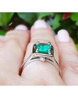 White gold ring with a natural emerald gemstone on a hand against a blurred green leafy background