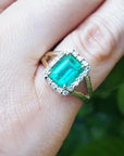An emerald-cut emerald ring on a hand with a natural background.