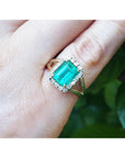 An emerald-cut emerald ring on a hand with a natural background.
