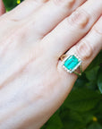 Hand wearing a natural emerald ring with a diamond halo against a green leafy background.