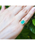 Hand wearing a natural emerald ring with a diamond halo against a green leafy background.