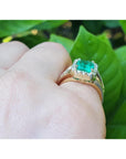 Emerald ring on a finger with a green leafy background.