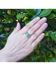Hand wearing a yellow gold mens solitaire ring with a real colombian emerald against a leafy background.