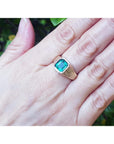 Hand wearing a ring with a square emerald against a blurred natural background.