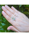 Hand wearing a pair of emerald earrings with geometric design against a green leafy background