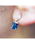 Gold earring with a blue sapphire and tapered baguette diamond worn on an ear