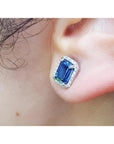 Emerald cut natural blue sapphire stud earring with diamond halo in 14K white gold on an ear.