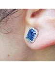 Emerald cut natural blue sapphire stud earring with diamond halo in 14K white gold on an ear.