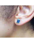 Emerald cut natural blue sapphire stud earring with diamond halo in 14K white gold on an ear.