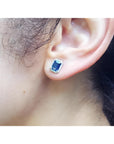 Emerald cut natural blue sapphire stud earring with diamond halo in 14K white gold on an ear.