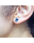 Emerald cut natural blue sapphire stud earring with diamond halo in 14K white gold on an ear.