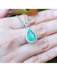 Green teardrop emerald pendant on a gold chain held on a hand with a blurred green background.