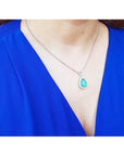 A woman wearing a blue top with a necklace featuring a teardrop emerald pendant.