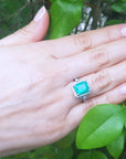 Hand wearing a Colombian emerald engagement ring with green leaves in the background.